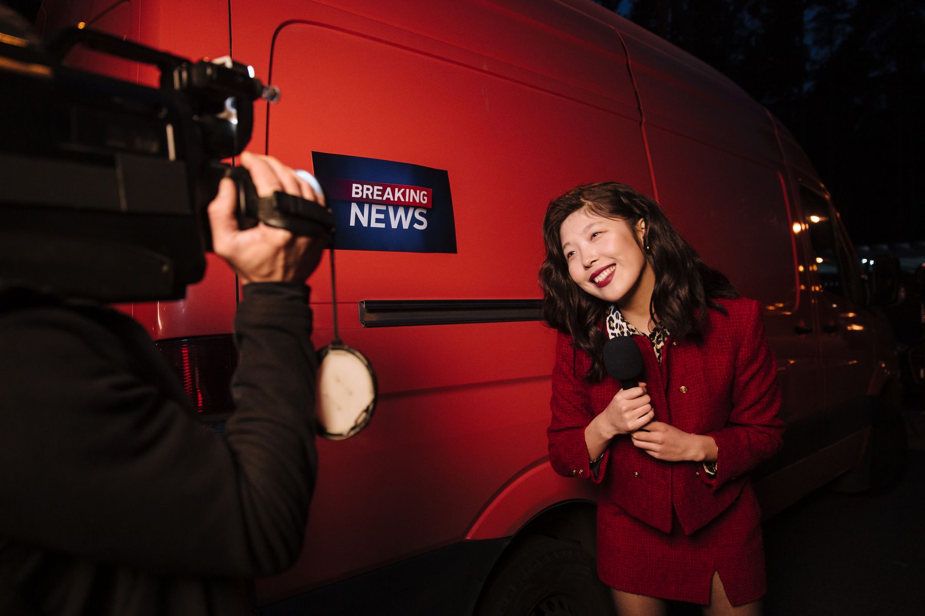 A smiling TV reporter holding a microphone in front of a news van while being filmed